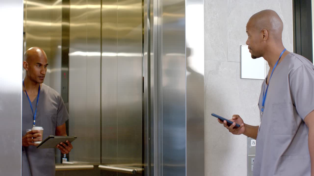 Dois funcionários masculinos do hospital a premir o botão do elevador com o telemóvel, enquanto um colega sai com um tablet e uma chávena, cumprimentando-os.