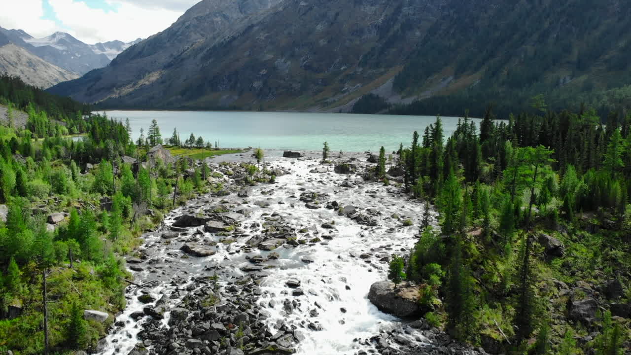 A breathtaking view of a mountain lake fed by a rushing river surrounded by lush forests and majestic peaks