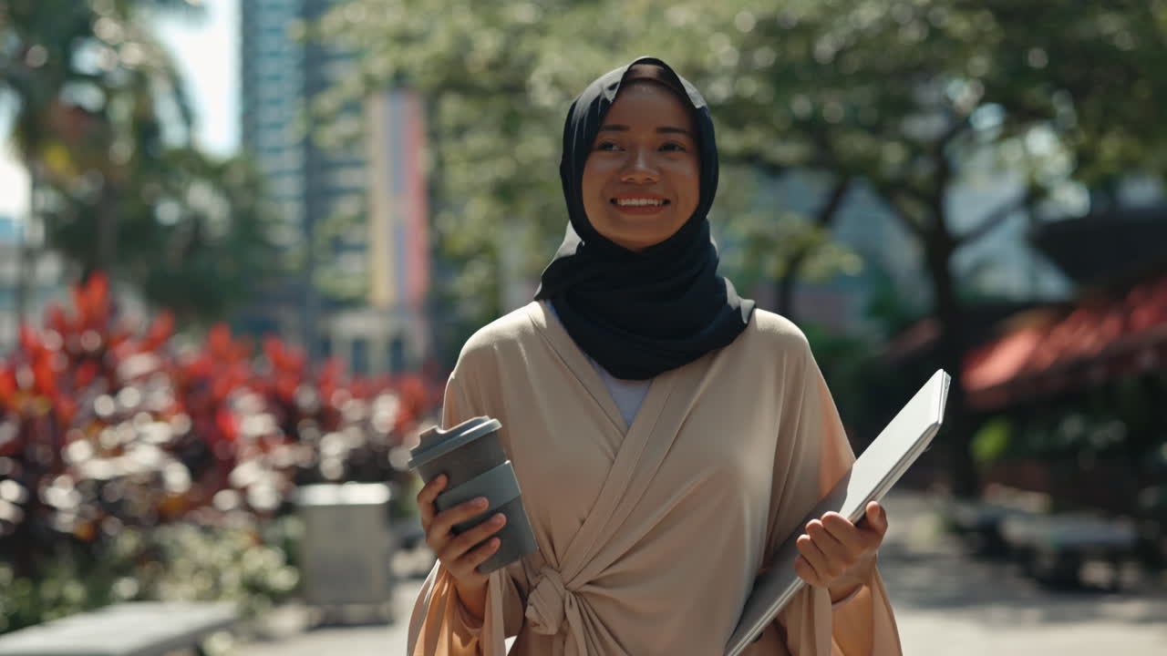 Smiling Young Muslim Woman in Hijab Walking Outdoors with Coffee and Laptop