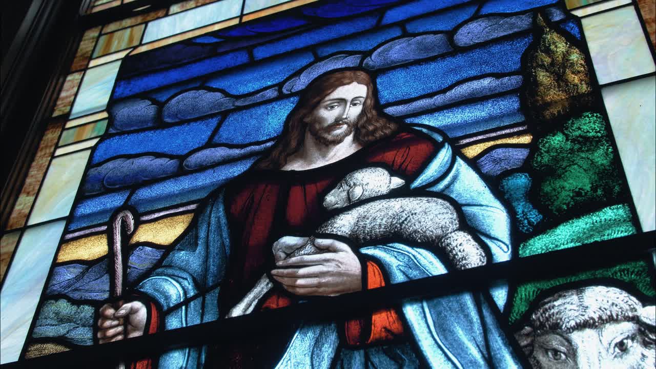 Vibrant stained-glass art inside a church sanctuary shows Jesus holding a lamb, surrounded by sheep, representing peace, faith, and religious tradition