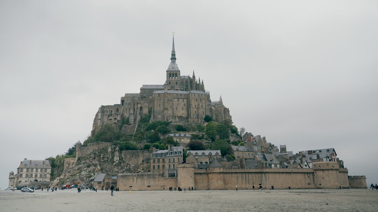 Mont St Michel - A massive, historic abbey sits atop a rocky island, surrounded by empty tidal flats under a cloudy sky, creating a striking contrast with the vast expanse of sand and sky.