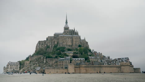 mont st michel - a massive, historic abbey sits atop a rocky island, surrounded by empty tidal flats under a cloudy sky, creating a striking contrast with the vast expanse of sand and sky.