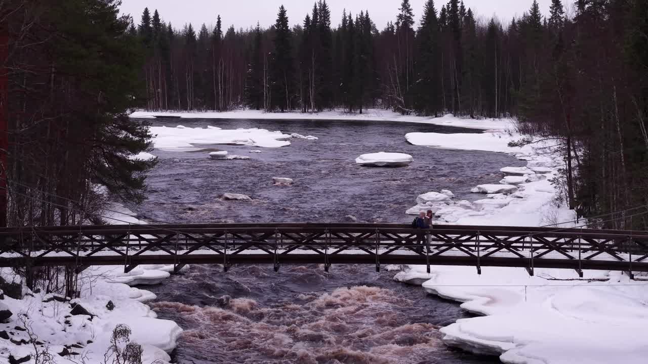 Pariskunta kävelee sillalla punertavan ruskean sävyisen joen yläpuolella, joka virtaa mäntymetsän läpi, Suomi, ilmakuva