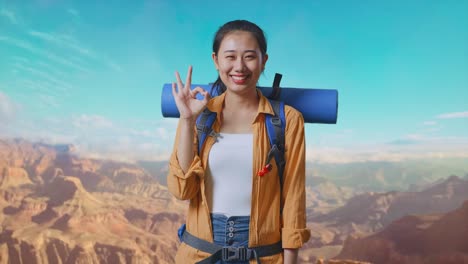 asian female hiker with mountaineering backpack smiling and showing okay gesture to camera while traveling at the top of mountain