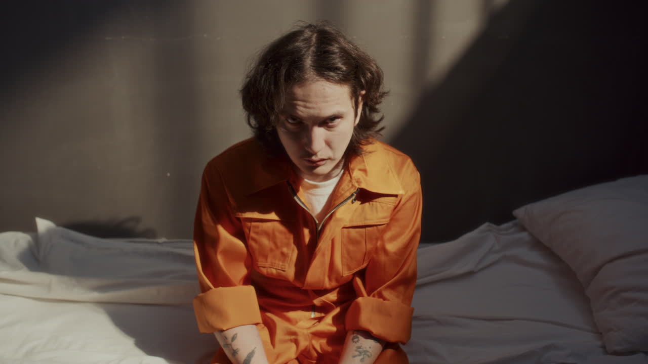 Portrait of Angry Young Man in Orange Uniform Sitting on Bed in Prison Cell