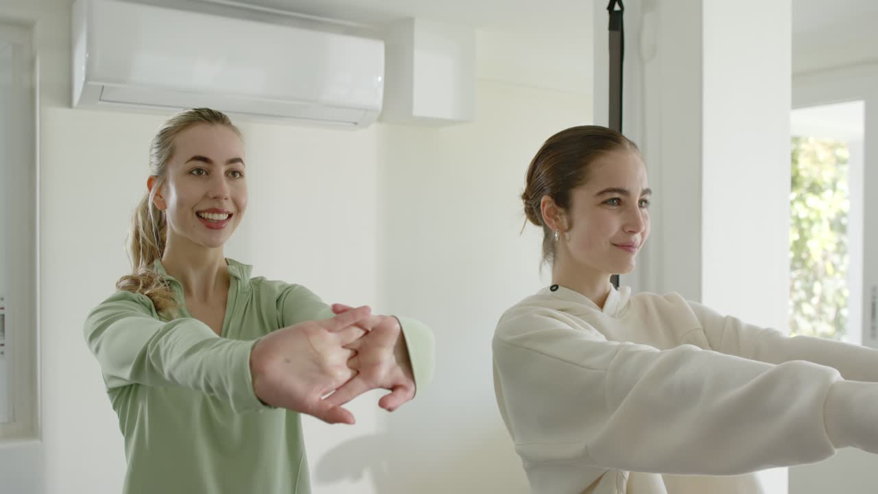 Female duo interlacing fingers, extending wrists in home gym under AC, band strap by foliage window