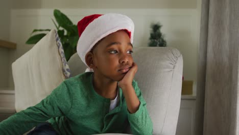 Trauriger-Afroamerikanischer-Junge-Mit-Weihnachtsmütze,-Der-Zur-Weihnachtszeit-Auf-Dem-Sofa-Sitzt