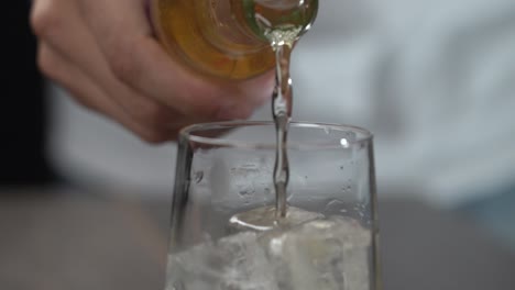 Making cocktail pouring ginger ale mixer from glass bottle into tall glass with ice close up on fizz