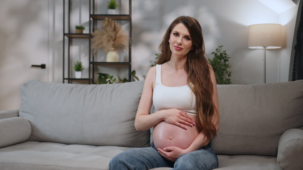 Young expectant mother sits comfortably on sofa cradling her prominent baby bump with gentle hands