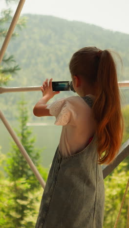 child takes video of lake and mountain at glamping. little girl holds videocamera by panoramic window at eco hotel. kid plays camerawoman at resort