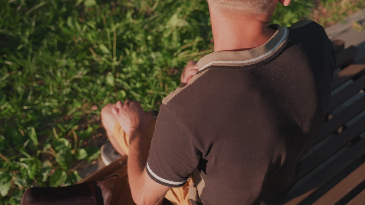 Young Man Sitting On Bench In Sunlit Grass, Rear Head And Shoulders Visible, Hands Resting On Knees, Close Rear View Captures Hairline, Collar And Wooden Seat Texture, Warm Summer Light And Quiet Park