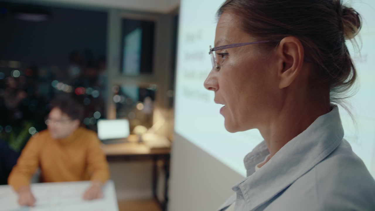 mujer de negocios experimentada de pie junto a la pantalla de proyección y respondiendo preguntas después de dar una presentación durante la reunión nocturna con colegas en la oficina. tiro en el pecho, centrarse en primer plano