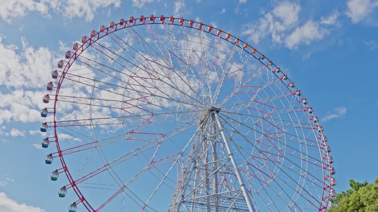 Expansive low-angle view showing the full height and structure of the large white and red Ferris wheel under a blue sky with clouds