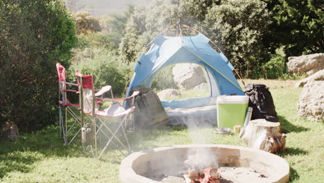 Rauchende-Feuerstelle,-Zelt,-Stühle-Und-Campingausrüstung-In-Sonniger-Landschaft,-Kopierraum,-Zeitlupe