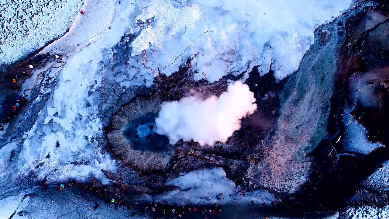 아이슬란드 게이시르에 있는 게이저 위의 공중 풍경: 많은 연기가 게이저 밖으로 나오고 있으며, 사람들이 해가 지는 곳에서 게이저를 돌아다니고 있다.