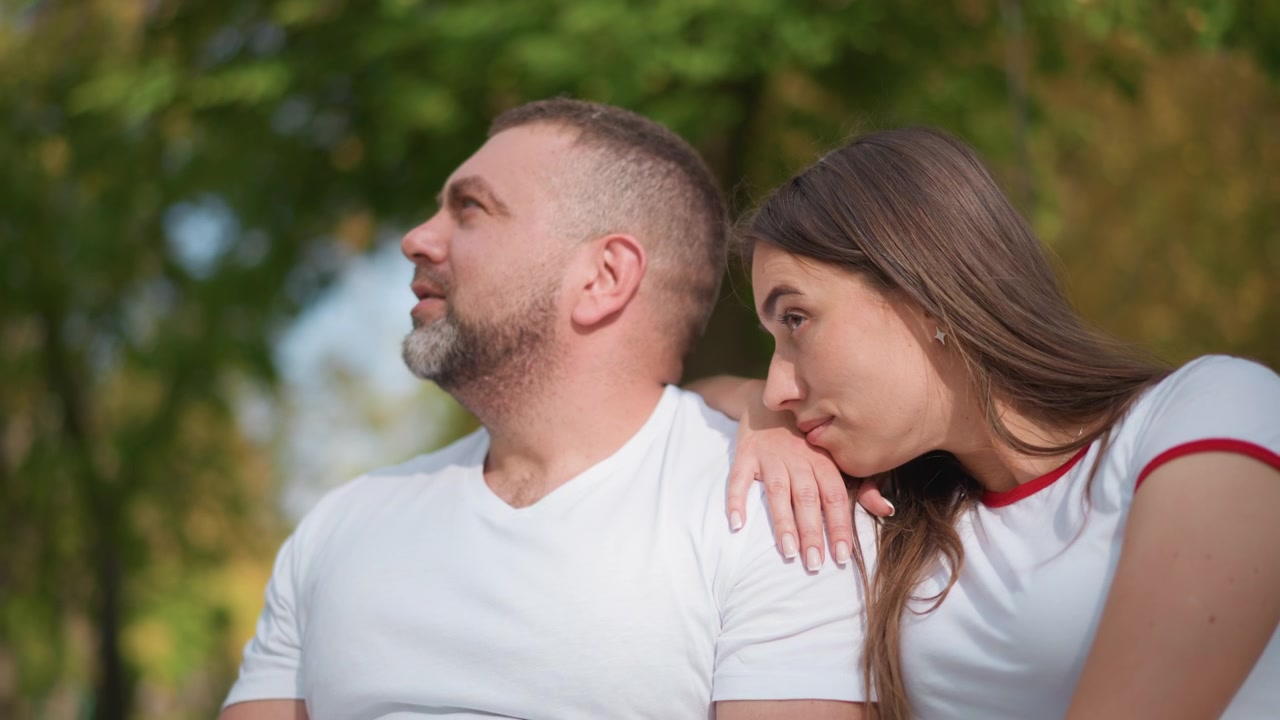 Primo piano di un uomo e una donna seduti insieme all'aperto, la giovane donna appoggia la testa sulla spalla di lui. Entrambi indossano camicie bianche con accenti rossi, una luce naturale soffusa illumina la loro postura rilassata.
