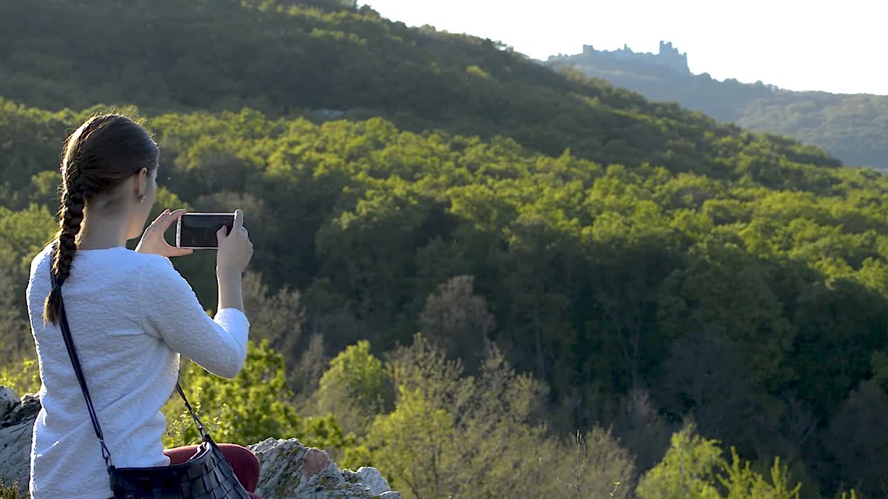 una joven con un suéter blanco y un pequeño bolso colgado del hombro tomando fotografías de las ruinas del antiguo fuerte en la distancia