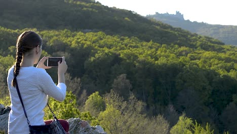 Niña-En-Un-Suéter-Blanco-Con-Un-Bolso-Pequeño-Colgando-Alrededor-De-Su-Hombro-Tomando-Fotos-De-Las-Antiguas-Ruinas-De-La-Fortaleza-En-La-Distancia