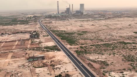 Cinematic-aerial-shot-of-silver-car-driving-on-country-road-in-desert-and-big-chimneys-in-distant-background