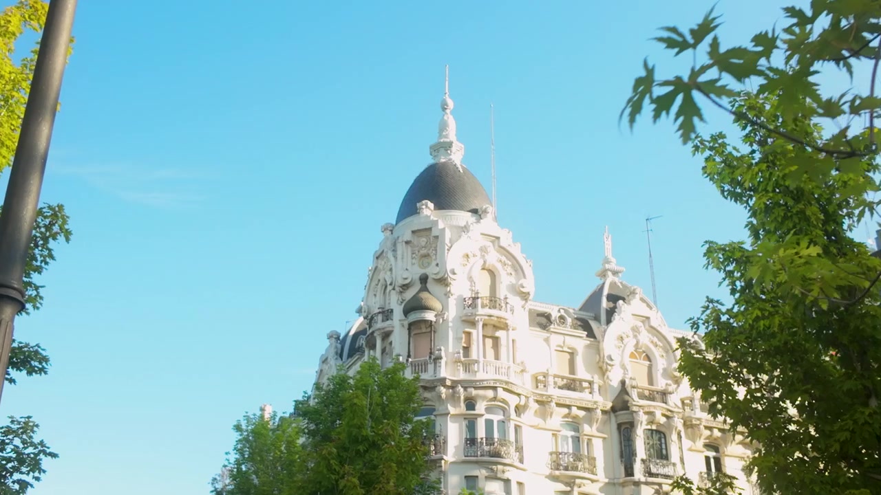 Un'inquadratura dal basso di edifici storici ornati a Madrid, Spagna, incorniciati da alberi verdi e rigogliosi sotto un cielo blu chiaro. Perfetto per documentari di viaggio e contenuti sullo stile di vita urbano.