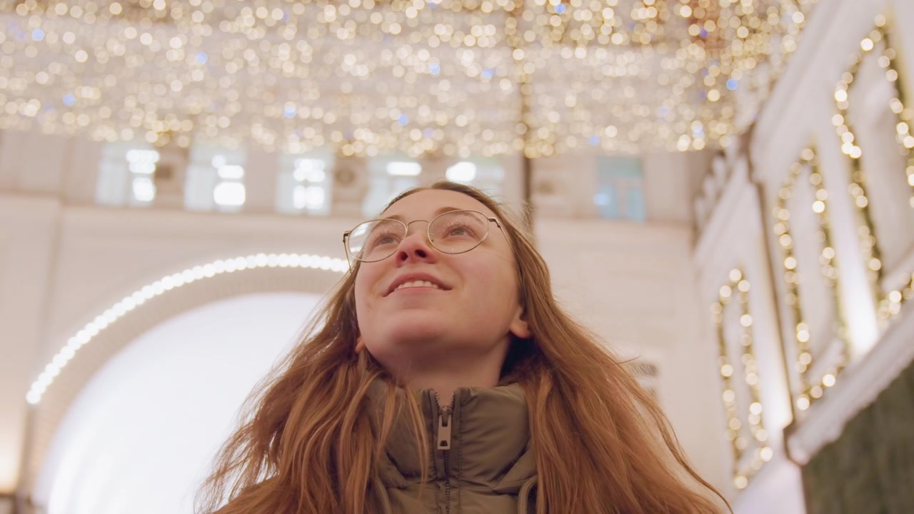 Giovane donna ammira le luci festive, con berretto accogliente e cappotto invernale in un'elegante galleria interna, decorazioni dorate sul soffitto e archi illuminati, lenta panoramica spontanea attraverso il corridoio dello shopping, calda