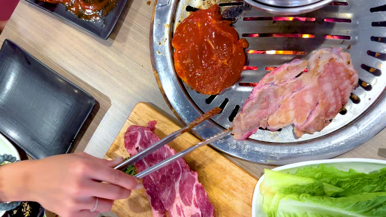 Hands grill marinated meat on a Korean BBQ setup in a vibrant Bangkok restaurant. Bright lighting highlights fresh ingredients and sizzling action
