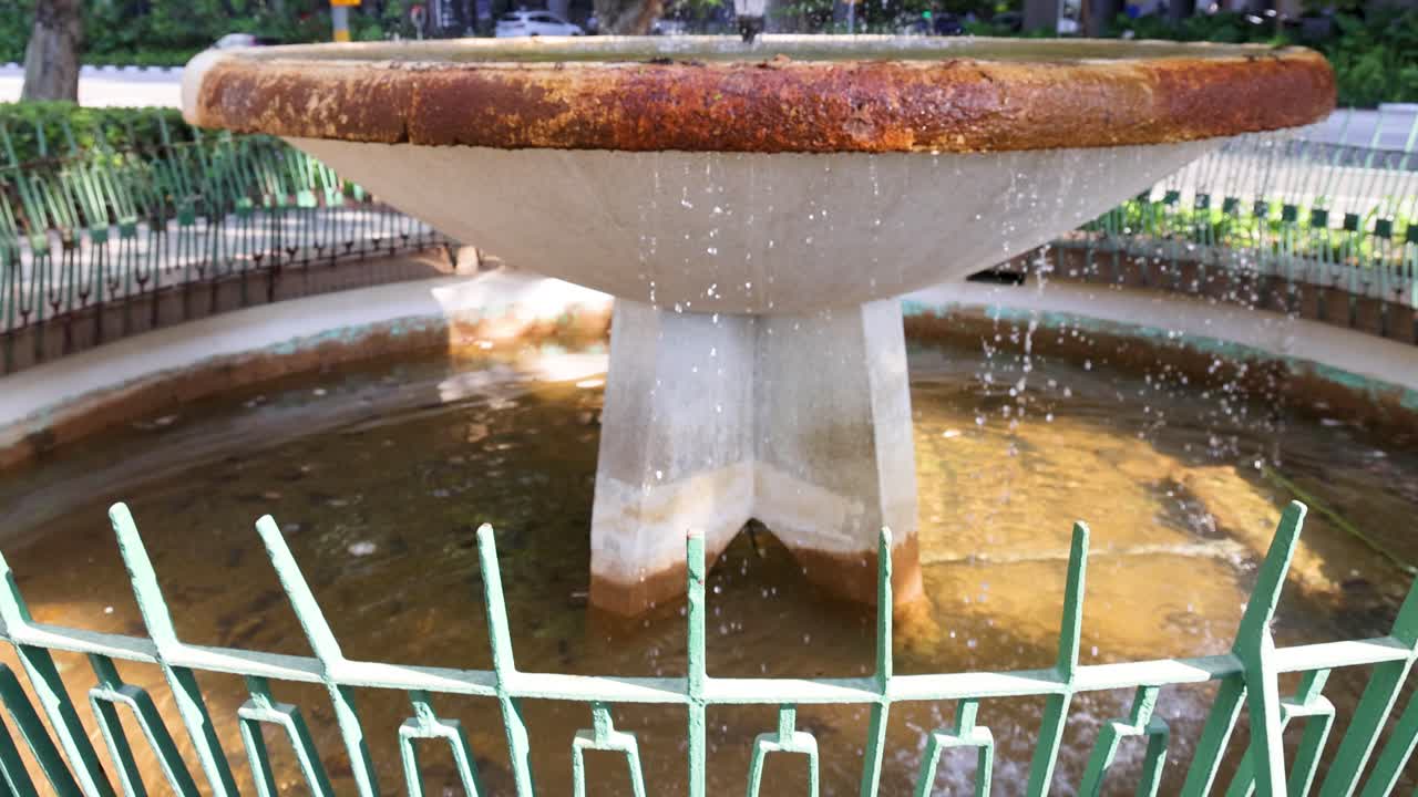Concrete fountain sprays water in landscaped city plaza, daylight, steady camera, lush greenery surrounds
