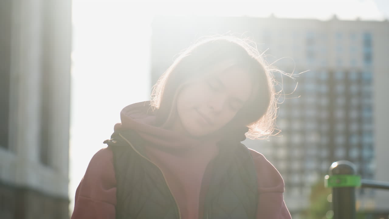 Fit female athlete with eyes closed stretches neck round while warming up outdoors against urban background with sunlight highlighting hair and serene concentration before training session routine