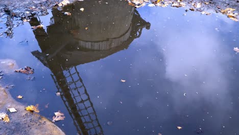 Altmodisches-Holzsegel-Vernachlässigte-Windmühlenreflexion-In-Wasserpfütze-Mit-Vorbeiziehenden-Wolken-Darüber