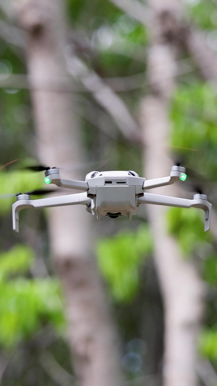 Bílý kvadrokoptérový dron se stabilně vznáší na rozmazaném pozadí stromů tropického lesa