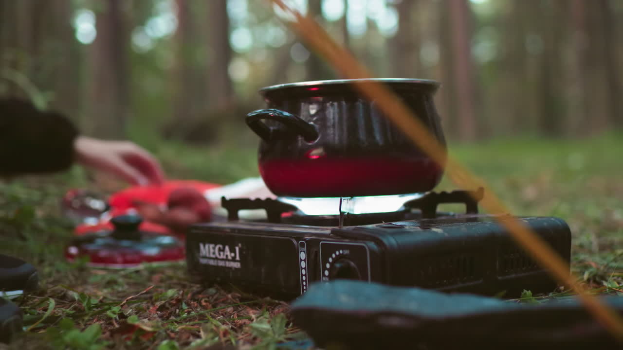 close up of lady turning on gas stove and picking knife to slice onion while companion pours water into black pot set on portable burner in forest setting surrounded by camping tools and cooking gear