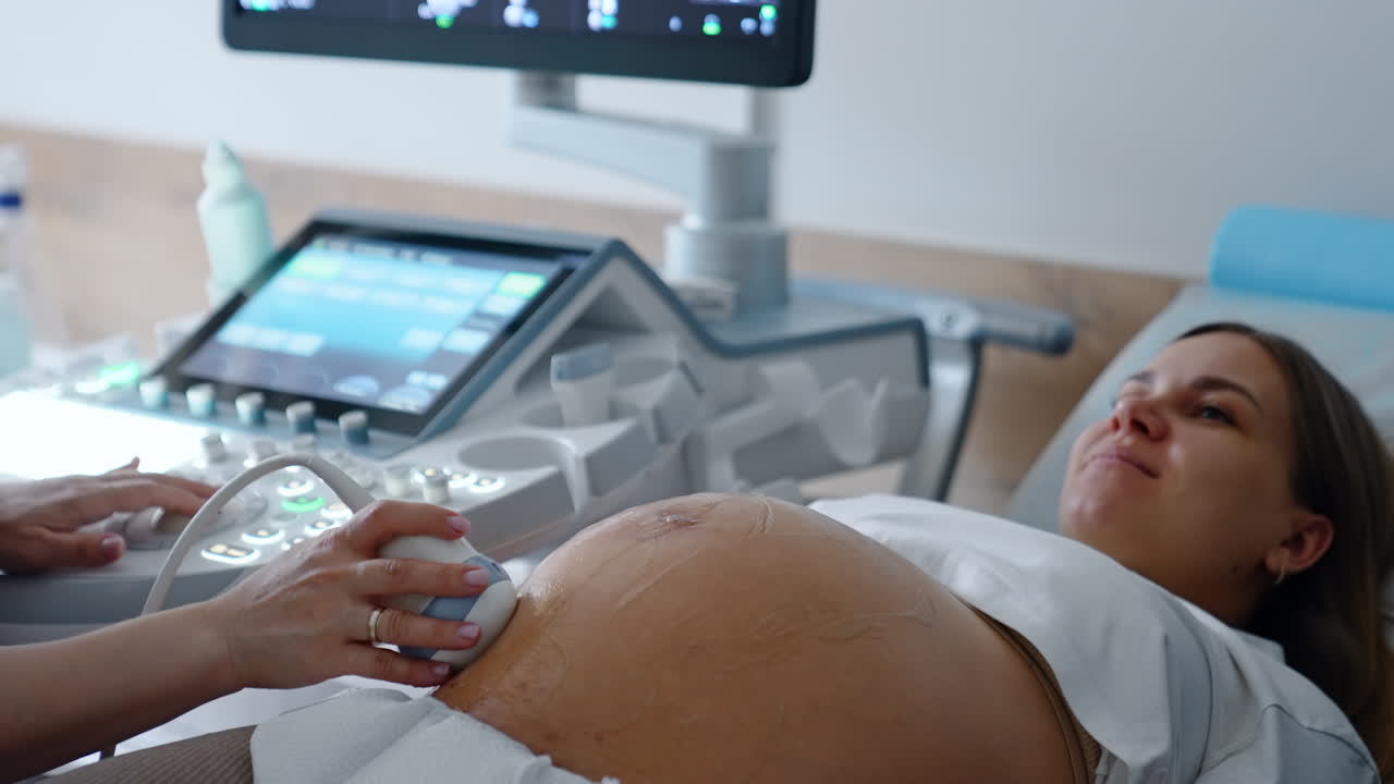 Expecting Caucasian woman having ultrasound diagnostics. Unrecognized female doctor moves the ultrasonic device by big round belly.