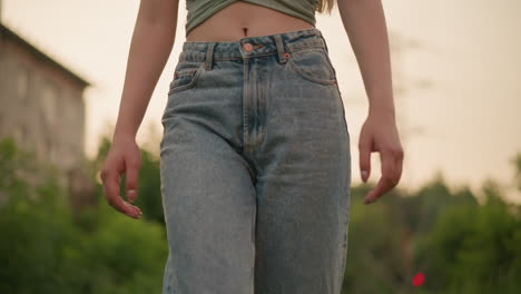 close-up of woman wearing high-waisted jeans and crop top, revealing her stomach, walking outdoors with blurred background of greenery, power lines, and urban structure