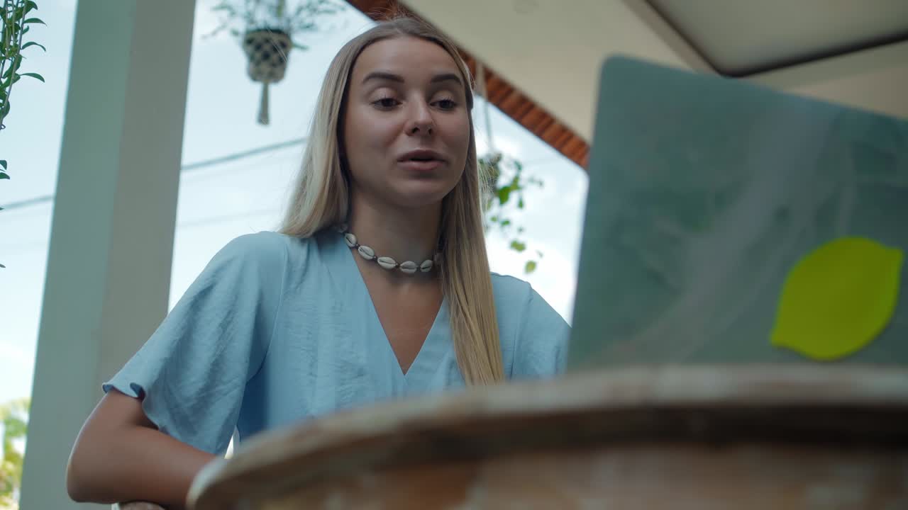 una chica en un vestido azul remotamente en línea trabajando detrás de una computadora portátil y mirando a la pantalla el patio trasero con plantas verdes