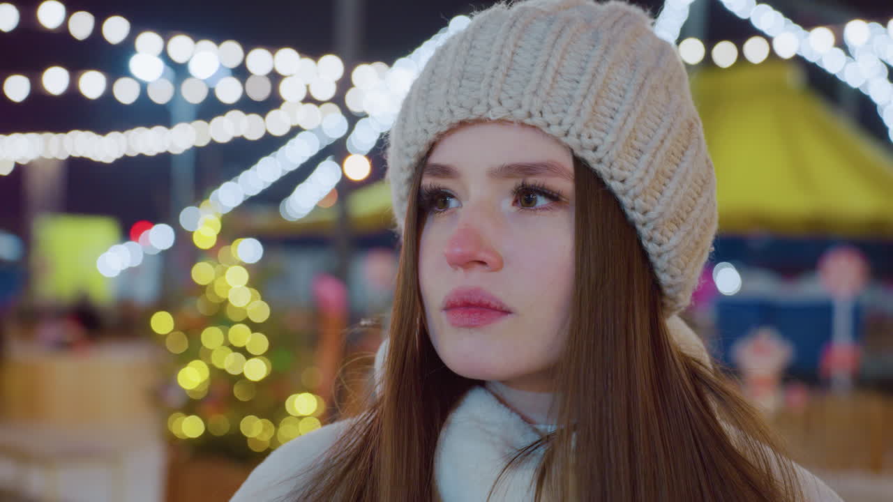 Close-up of woman wearing cozy beanie and winter coat, gazing thoughtfully with a contemplative expression, soft festive lights in background
