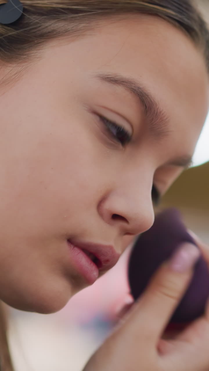 primer plano de una adolescente aplicándose corrector con una esponja, expresión concentrada, fondo bokeh suave, iluminación interior de centro comercial, difuminado preciso bajo los ojos con una esponja morada, textura de piel natural, autocuidado tranquilo