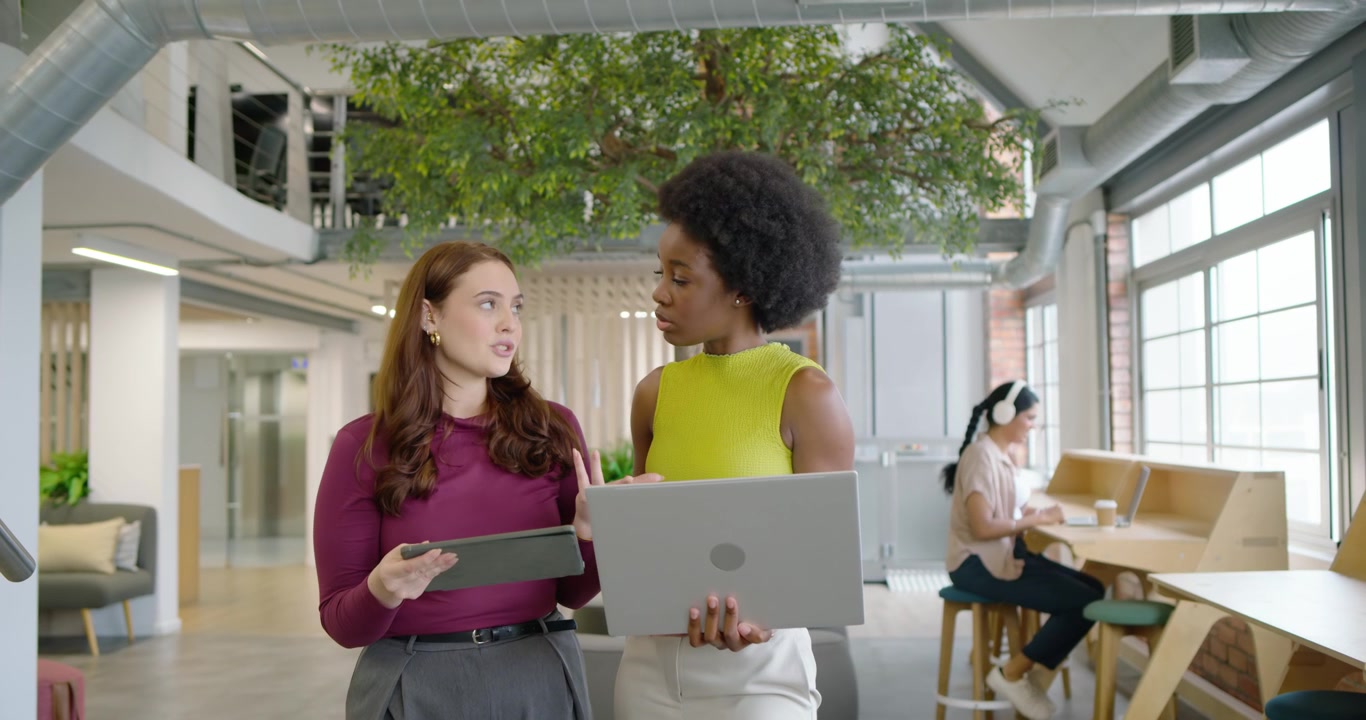 Diverse women team working on tablet laptop in office as peer approaching with coffee joining chat