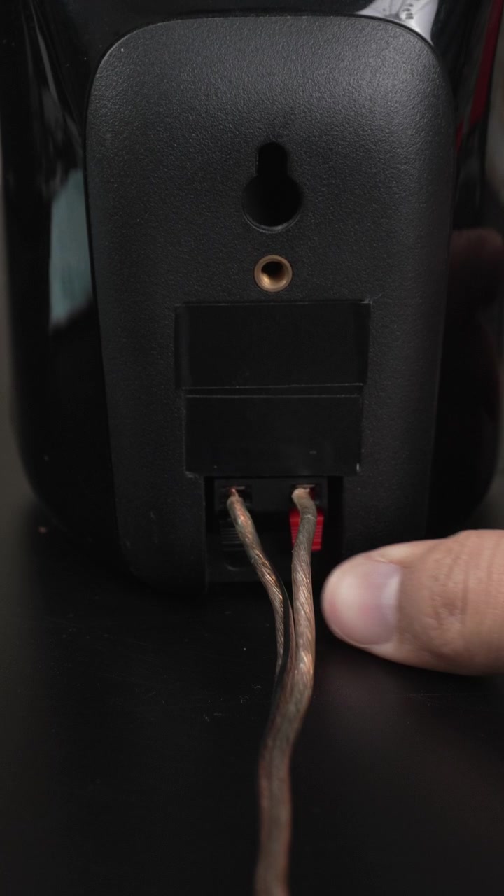 Hands attach bare speaker wire to the spring clip terminals on the back of a black bookshelf speaker on a desk, useful for tutorials, product demos, or home o guides.