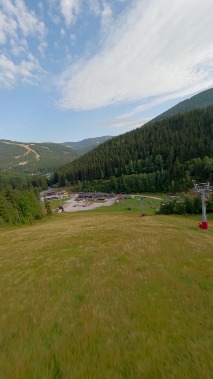 Ripresa aerea verticale dinamica FPV che sorvola un lussureggiante resort di montagna verde con impianti di risalita e pinete sotto un cielo nuvoloso. Perfetto per contenuti di viaggio e avventura all'aperto