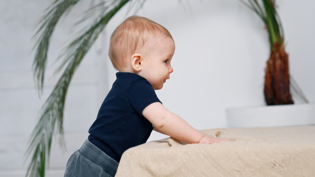 Red-haired infant boy standing indoors. Cute little baby holding by the sofa smiling adorably. Side view.