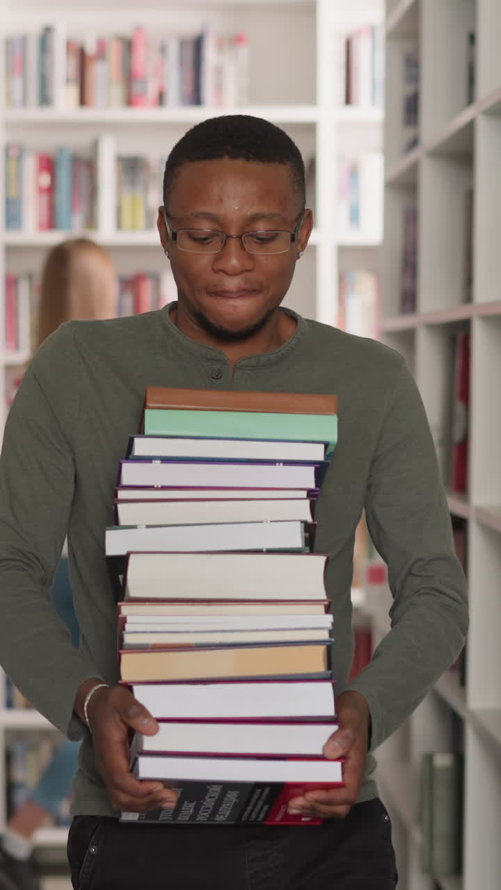 Black man with book stack in library. African American customer carries large literature pile walking along aisle between bookcases. Information learning