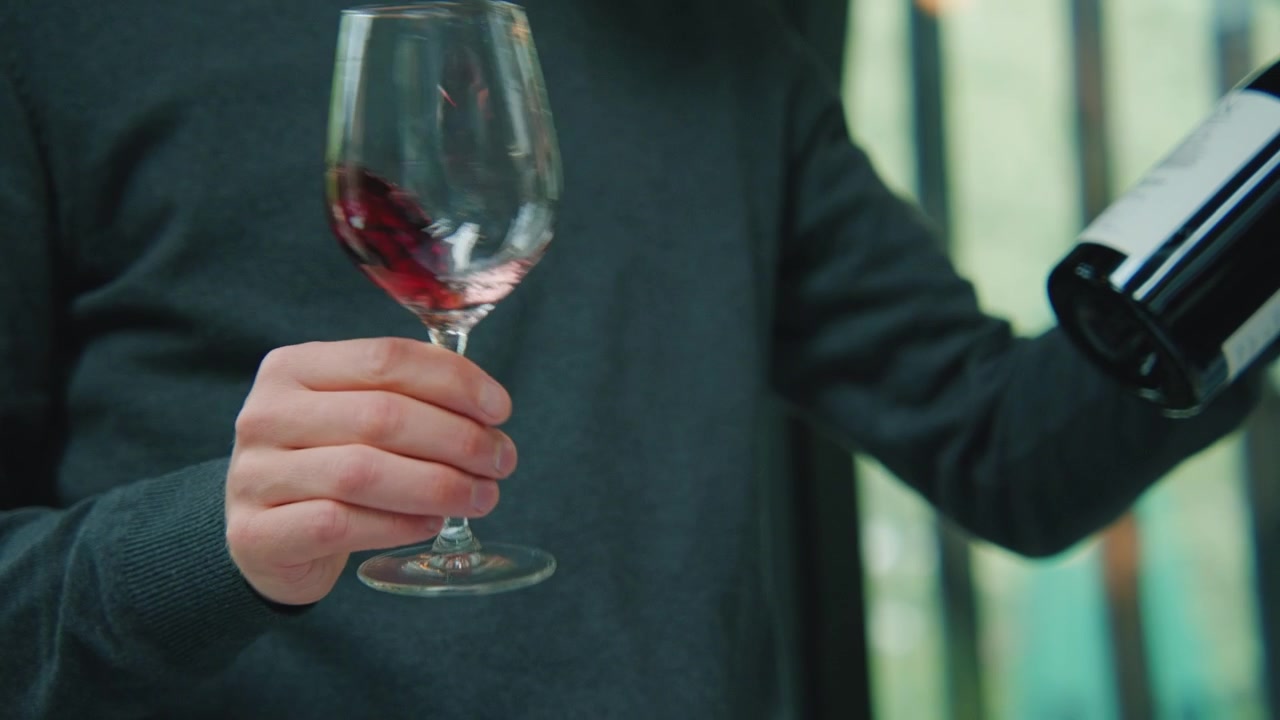Un hombre sosteniendo una botella de vino y una copa llena con un poco de este, agita la copa con vino antes de probarlo