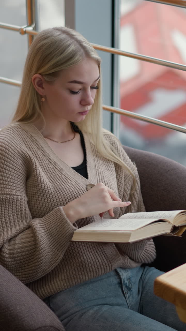 una mujer pensativa pasa a una nueva página mientras lee un libro en un acogedor café con vista a la ciudad a través de un panel de vidrio, una taza de café y un teléfono colocados en la mesa delante de ella