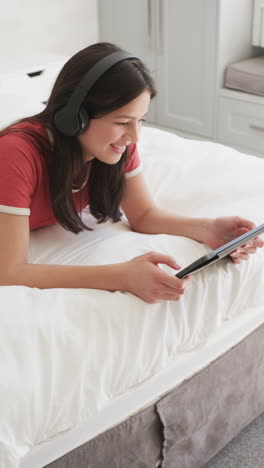 vertical video of happy biracial woman in headphones lying on bed using tablet, slow motion