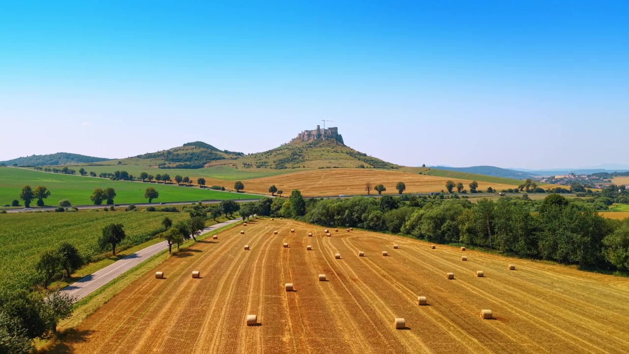 Paesaggio aereo di una strada vicino a un campo mietuto e alla collina del Castello di Spiš. Prospettiva dall'alto di una strada asfaltata che costeggia un campo giallo con balle di fieno rotonde, alberi verdi e un castello sullo sfondo.
