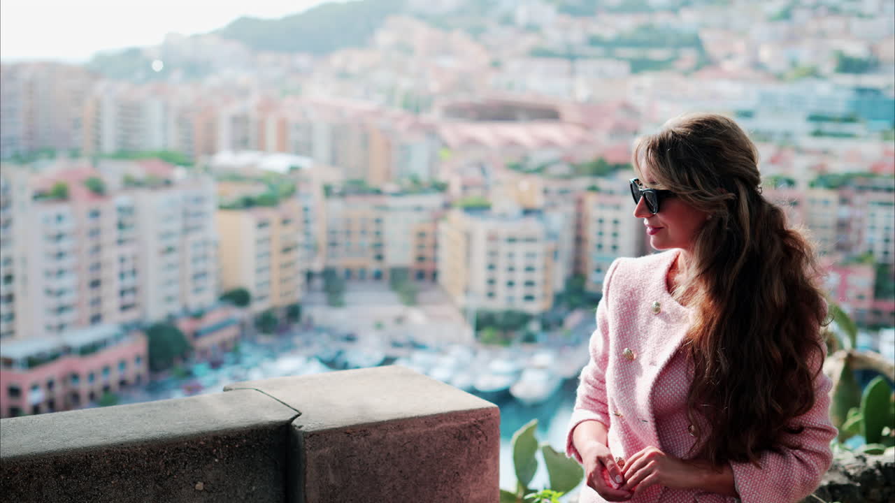 Brunette woman in a pink blazer smiling with a blurry view of boats docked in the Port de Fontvieille in Monaco