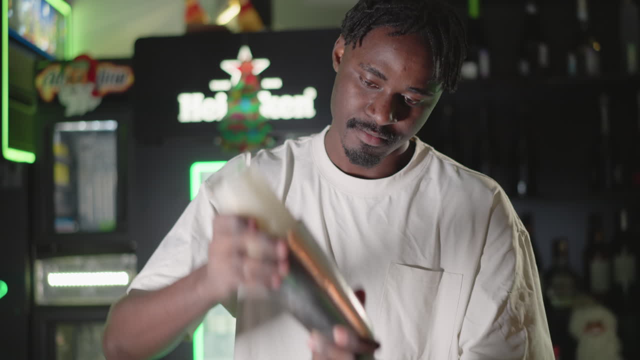Bartender shaking cocktail shaker with focus on hands in professional bar setting. Behind, illuminated liquor shelves and Heineken neon sign glowing brightly, setting the nightlife scene