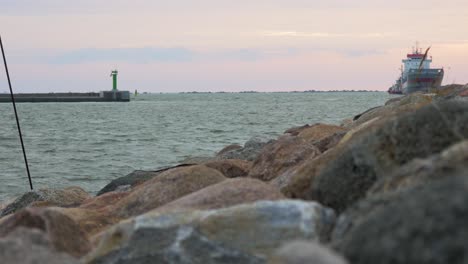 Large-gray-cargo-ship-entering-the-Port-of-Liepaja-in-calm-sunny-summer-day,-stone-pier-in-foreground,-waves-splashing,-distant-medium-low-angle-shot