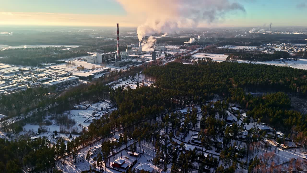 Vinterpanorama af et industrielt kraftværk med skorstene, der rager op over en sneklædt skov og en nærliggende bebyggelse.