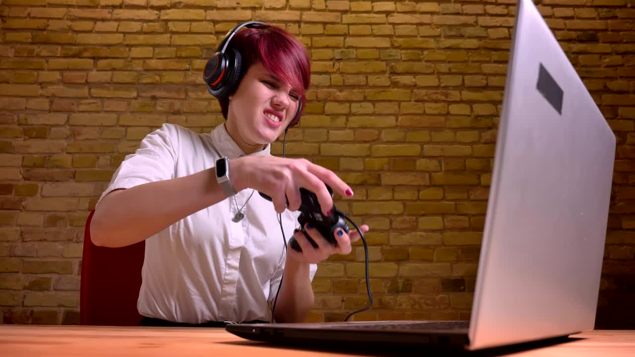 짧은 머리카락의 소녀 스트리머는 매니큐어와 헤드폰을 착용하여 벽돌 벽 바탕에 조이스틱을 감정적으로 연주합니다.
